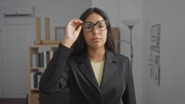 Genç esmer kadın, arka planda kitap rafları ve belgelerle bir ofis ortamında gözlük ayarlıyor.