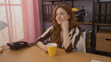 Çizgili tişörtlü, genç ve çekici bir sarışın kadın elinde sarı bir bardakla arka planda raflar ve dosyalarla rahat bir ofis masasında oturuyor..