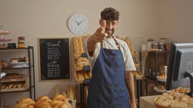 Önlüklü genç adam gülümsüyor ve ekmek ve hamur işleri sergilenen fırının içinde parmağıyla işaret ediyor, karatahta menüsü ve duvarda bir saat var.