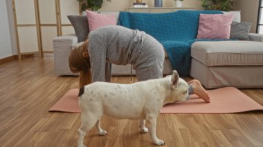 Genç sarışın bir kadın, evcil köpeğiyle birlikte oturma odasında yoga yapıyor..