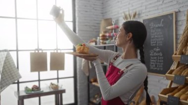 Fırında hamur işi ile selfie çeken bir kadın, ev ortamında taze pişmiş yiyecekler sergiliyor, kırmızı önlük ve beyaz gömlek giyiyor, neşeli bir atmosfer.