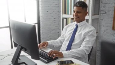 Beyaz gömlekli ve mavi kravatlı genç adam masasında bilgisayar ve akıllı telefon kullanırken tuğla duvar ve kitaplıklarla modern bir ofiste sohbet ediyor..