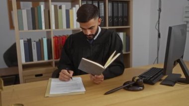 Yargıç cüppeli genç adam iş yerinde hukuk kitapları ve tokmakla ilgili belgeler okuyor ve imzalıyor.