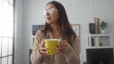Ofiste kahve içen bir kadın, arka planında sarı bir kupa ve kitaplık olan, gündelik ve modern bir iç mimariye sahip bir çalışma ortamı sergiliyor..