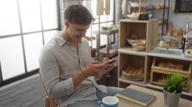 Genç adam, arka planda çeşitli hamur işleri ve ekmeklerle dolu bir fırında akıllı telefon kullanırken gülümsüyor.