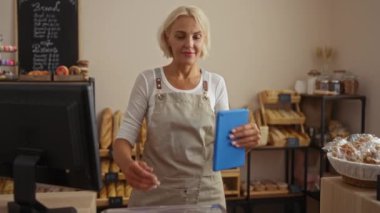 Fırında gülümseyen ve elinde tablet tutan bir kadın, çeşitli ekmek ve hamur işleriyle çevrili, kapalı bir dükkanda elinde bilgisayarla tezgahın arkasında duruyor.