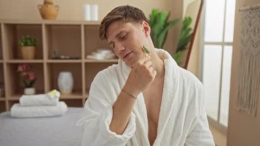 Genç adam, yumuşak ışıklandırmalı ve zarif dekorlu rahatlatıcı bir kaplıca ortamında yeşim taşıyla yüz bakımı yapıyor.