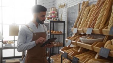 Fırındaki genç adam önlük giyerken tablet kullanıyor. Etrafı fırın ve raflarla çevrili.
