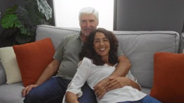 Orta yaşlı çift rahat bir oturma odasında rahat rahat oturuyor. Kadın ve erkek kanepede gülümsüyor ve evde ailecek vakit geçiriyorlar..