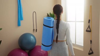 Spor salonunda elinde mavi yoga minderi tutan esmer bir kadın spor giysisi giyiyor. Arka planda spor topları ve spor aletleri var.