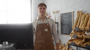 Önlük giyen genç adam, arka planda bir karatahta menüsü ve ekmek rafları olan bir fırının içinde güvenle duruyor.