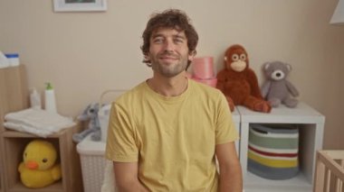Doldurulmuş oyuncaklar ve bebek eşyalarıyla dolu rahat bir yatak odasında gülümseyen genç bir adam. Parlak ve hoş bir ev ortamında rahat ve neşeli bir atmosfer sergiliyor..