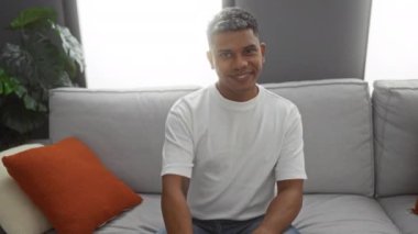 Genç adam, rahat bir oturma odasında, yastıklar ve bitkilerle çevrili, rahat ve misafirperver bir iç atmosfer sergileyen, samimi bir gülümsemeyle kamerayı işaret ediyor..