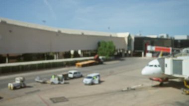 Bulanık havaalanı sahnesi, kapalı araçlar, işçiler ve güneşli bir günde arka planda bir uçak ile asfalt aktivitelerini gösteriyor.