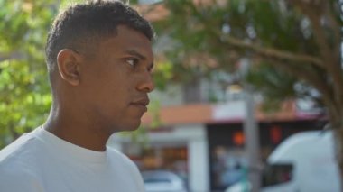 Arka planda ağaçlar ve binalar olan şehir parkında beyaz tişörtlü genç İspanyol bir adam.