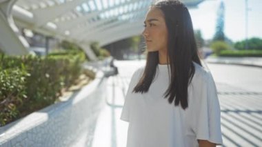 Şehir parkında modern mimarisi olan genç bir kadın beyaz bir gömlek giyiyor ve güneşli bir günde kameraya gülümsüyor.