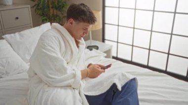Bornozlu genç adam, modern pencereli ve minimalist dekorlu parlak bir yatak odasında yatarken akıllı telefon kullanıyor.