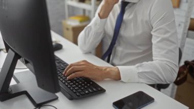 Ofis masasında oturan adam yorgun görünüyor. Gömlek ve kravat giyiyor. Etrafı bilgisayar, kitap ve telefonla çevrili..