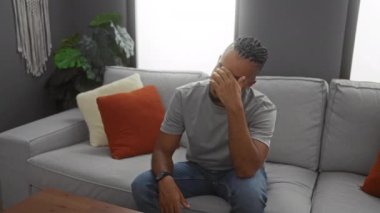 Modern bir oturma odasındaki kanepede oturan genç adam şık dekorlar ve canlı yastıklarla çevrili düşünceli bir yüz ifadesi sergiliyor..