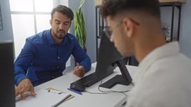 Parlak bir ofiste birlikte çalışan iki adam bilgisayarı, bitkileri ve arka planda görünen bir strateji tahtası olan bir masada belgeleri tartışıyorlar.