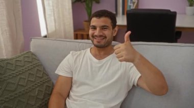 Genç adam apartmanın oturma odasında baş parmak hareketi yapıyor.