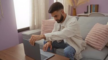 Genç İspanyol bir adam, rahat bir oturma odasında dizüstü bilgisayarıyla çalışıyor. Yumuşak pembe dekorlu ve rahat gri bir kanepesi var. Tahta bir kahve masasında otururken ekranına odaklanmış..