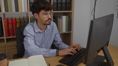 Ofis odasındaki bilgisayarda çalışan genç bir adam. Yanında kitap rafları ve kahve fincanı var.