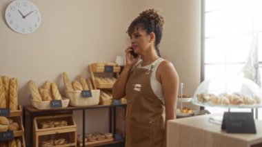 Fırında telefonla konuşan bir kadın, ekmek vitrinlerinin ve pasta raflarının yanında duruyor, iç mekan ayarları, önlük giyiyor, kıvırcık saçlı, gülümseyen, iç mekan aydınlatması, İspanyol, kadın, yetişkin