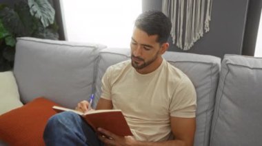 Genç bir adam evinde rahat bir oturma odasında rahat bir koltukta deftere yazı yazıyor.