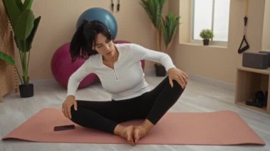 Spor salonunda yoga minderinde uzanan bir kadın. Etrafı bitkilerle, egzersiz toplarıyla ve spor aletleriyle çevrili.