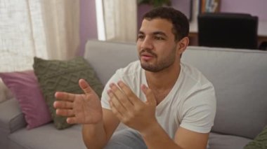 Genç adam oturma odasında dinleniyor, gülümsüyor ve ışık perdeleri ve rahat kanepe yastıklarıyla ev ortamının keyfini çıkarıyor..