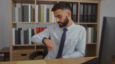 Ofiste sakallı ve kravatlı genç bir adam arka planda kitaplıkla masa başında otururken kol saatine bakıyor.