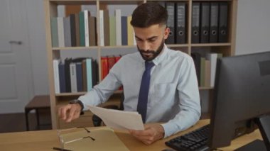 Genç İspanyol adam iş yerindeki belgeleri inceliyor. İçeride rafları ve bilgisayarı var.