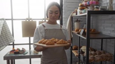 Pastanedeki genç İspanyol kadın elinde kruvasan tepsisiyle evin içindeki hamur işleri raflarının yanında gülümsüyor.