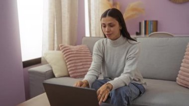 Genç bir kadın rahat bir oturma odasında dizüstü bilgisayar kullanıyor. Düşünceli, şaşkın ve evde çeşitli duygusal tepkiler sergiliyor..