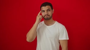 Beyaz tişörtlü genç İspanyol adam parmak kaldırarak ayakta duruyor, başarılı bir fikirle mutlu; bir numaralı hareket izole edilmiş kırmızı arka plan üzerinde