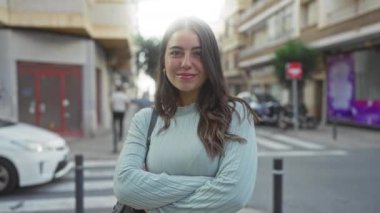 Esmer saçlı bir kadın şehir sokağında güven içinde duruyor, kollarını kavuşturmuş, arka planda arabalar ve binalar var, güzel ve sofistike bir şehir..