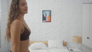 Genç, İspanyol, mor spor sutyenli bir kadın günışığında modern, beyaz tuğlalı bir yatak odası, tropikal bitki ve soyut sanatla eğleniyor ve poz veriyor.