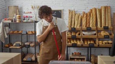 Genç adam arka planda ekmek rafları ve hamur işleriyle fırında süpürge sapına şarkı söylüyor.