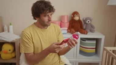 Yatak odasındaki genç adam, oyuncaklarla ve bebek eşyalarıyla telefonla şişeyi inceliyor, ebeveynlikte düşünceli bir an yaşıyor..