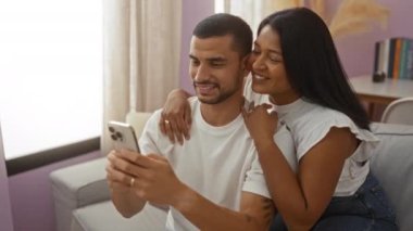 İspanyol çift evlerinde rahat oturma odalarında akıllı bir telefona bakarken mutlu bir şekilde kucaklaşıyorlar.