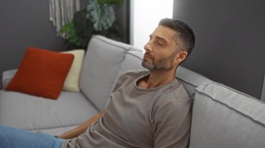 Genç adam modern bir kanepede dinleniyor rahat bir oturma odasında şık dekorlu ve nötr renklerle, evdeki sakin ve iç gözlemci atmosferi iletiyor..