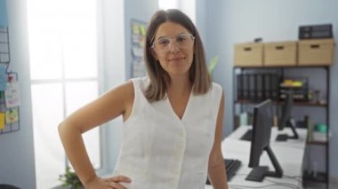 Sıraları ve arka planda rafları düzenleyen modern ofis ortamında gülümseyen ve bardakları kaldıran İspanyol kadın.