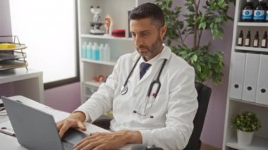 Klinikteki İspanyol doktor dizüstü bilgisayarla çalışırken boynunu tutuyor. Ağrı ya da rahatsızlık belirtileri gösteriyor. Etrafı tıbbi ekipmanlarla ve düzenli çalışma alanlarıyla çevrili..