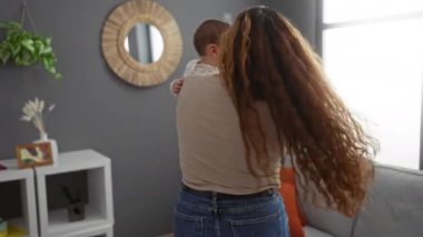 Anne, sıcak bir oturma odasında bebeğiyle neşeyle dans ediyor. Aşkı ve aile birlikteliğini yansıtıyor. Ev dekorunun ve doğal ışığın arka planına karşı..