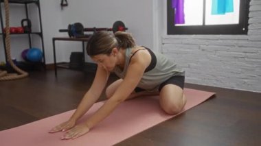 Spor salonundaki yoga minderinde gerinen ve arka planda spor ekipmanları olan bir kadın egzersiz pozu veriyor.