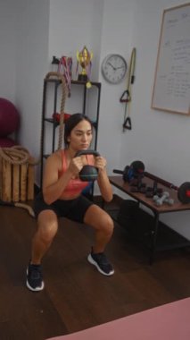 Spor salonunda kettlebell ile egzersiz yapan bir kadın. Etrafı spor aletleri ve ödüllerle çevrili. Odaklanma ve kararlılık öneriyor..