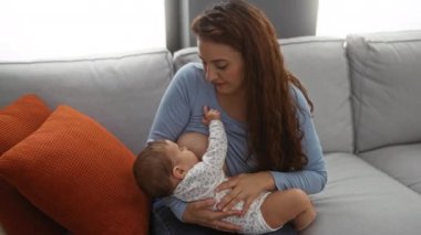 Bebek emziren bir kadın, rahat bir oturma odasında, turuncu yastıklarla, sıcak aydınlatmayla çevrili, sevgi ve şefkat ile kapalı bir evde aile anını gözler önüne seriyor..