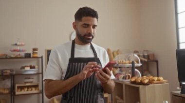 Kapalı bir fırın ortamında akıllı telefon kullanan genç adam. Etrafı fırın ve vitrinlerle çevrili.