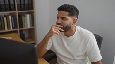 Ofis masasında çalışan genç bir adam, odaklanmış ve meşgul, arka planda kitaplıklar olan modern bir işyeri ortamında.